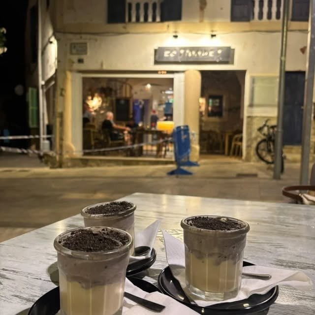 Vasos de café con crema y galleta triturada en una mesa de la terraza, de noche, con el local iluminado al fondo
